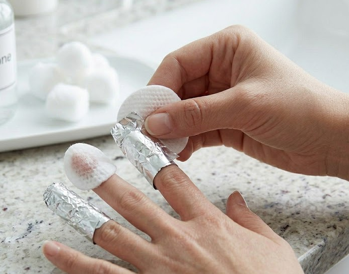 A close-up image showing a fingernail covered with an acetone-soaked cotton pad and tightly wrapped in aluminum foil to secure it.