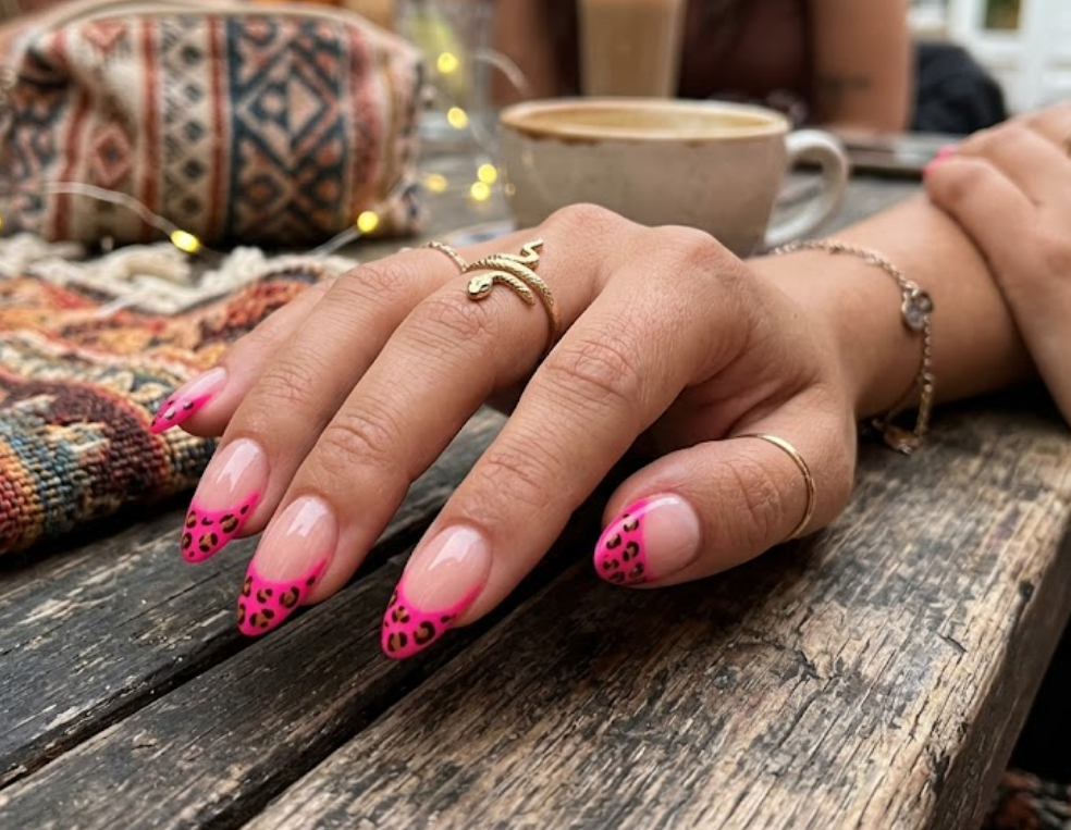 Realistic close-up of almond-shaped nails featuring neon pink French tips with leopard print spots, styled with gold rings on a rustic wooden table.