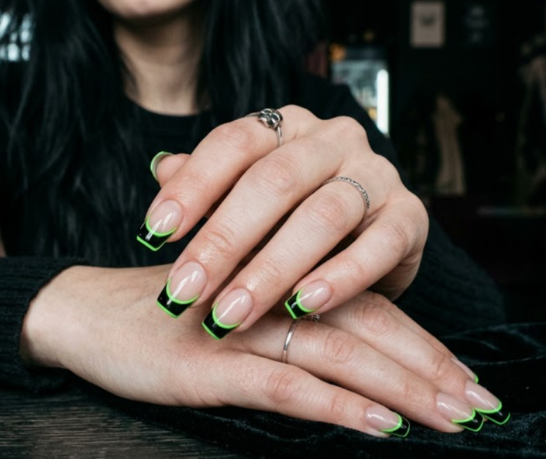 A close-up photo of a hand featuring jet-black French tip nails, each accented with a thin, vibrant neon green outline.