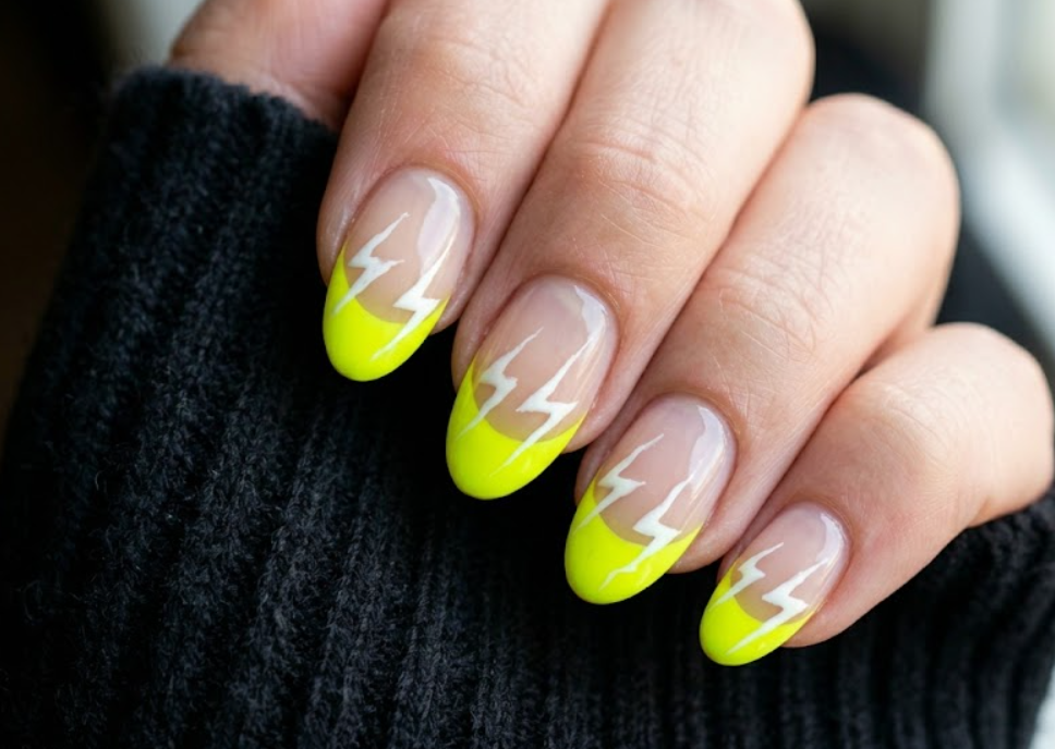 A close-up photo of a hand with oval-shaped nails featuring a natural base and bright neon yellow French tips. Each nail is accented with a sharp, jagged white lightning bolt design striking across the yellow tips.