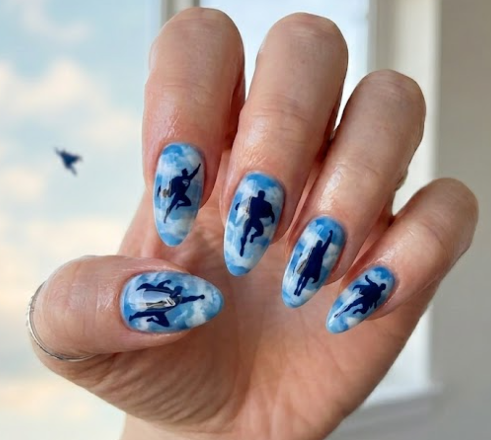 A woman’s hand with light blue nails, soft clouds, and small Superman silhouettes in a glossy finish.