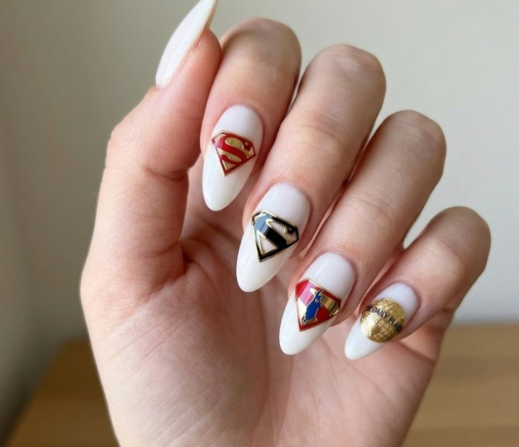 A woman’s hand with white base nails and small Superman “S” symbols placed on each nail in a minimal style.