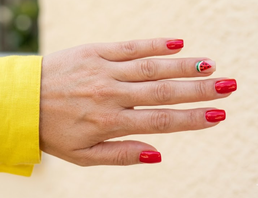 This image shows red summer nails featuring a watermelon accent design on a female hand.