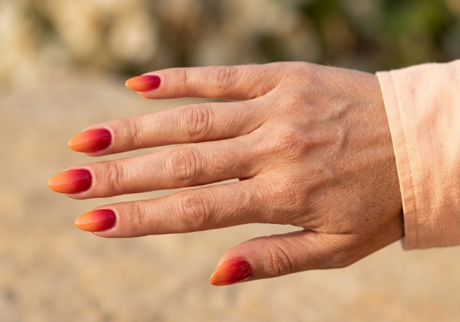 This image shows red-to-orange sunset ombre nails on a female hand with a peach summer sleeve.