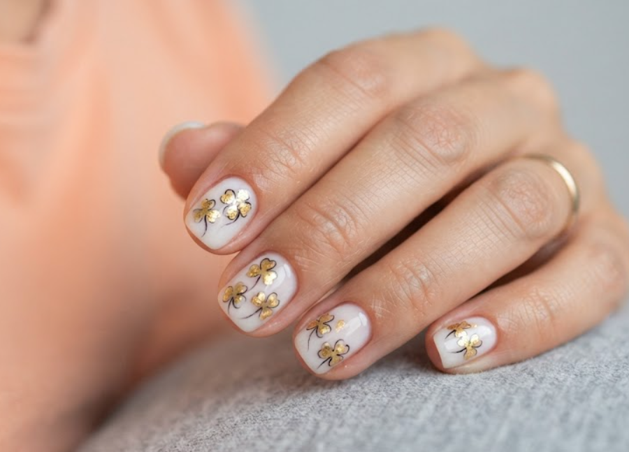 imageIn this image, clean nails feature a milky white base and gold foil clover designs while wearing a peach shirt.