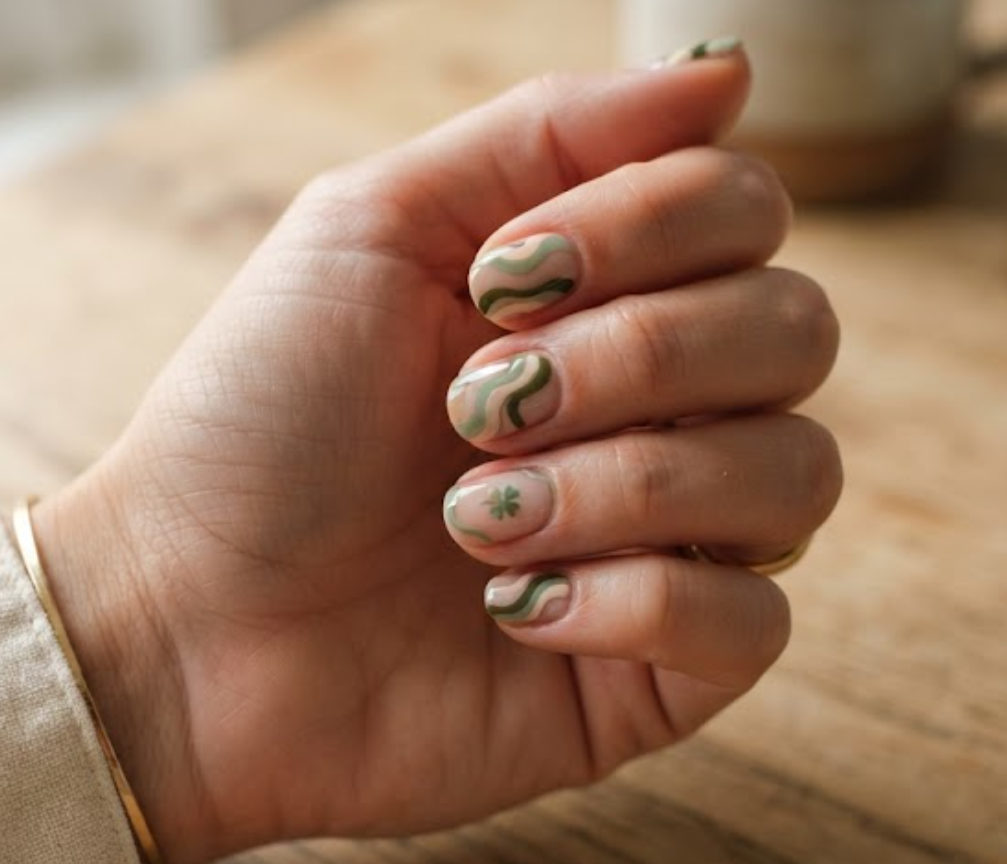 In this image, clean nails display retro sage green swirls and a minimalist clover while wearing a cream shirt.