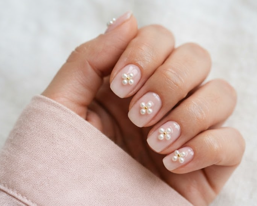 In this image, clean nails feature white pearls arranged into clover shapes while wearing a soft blush shirt.