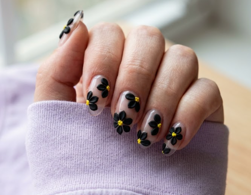 In this image, full hand nails feature tactile 3D sculpted black acrylic flower petals rising off a clean clear base, cradling tiny yellow/amber centers.