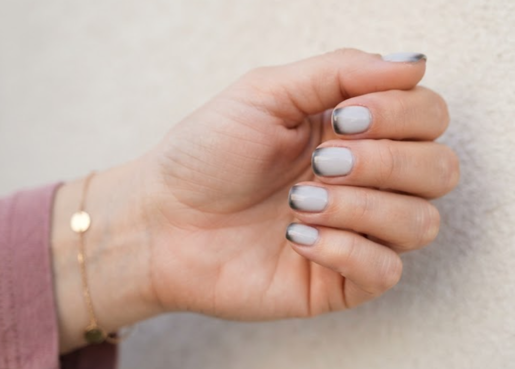 In this image, a full hand with extremely short squoval nails displays business nails with an ice-grey base and a smudged tip, wearing a dusty rose shirt.