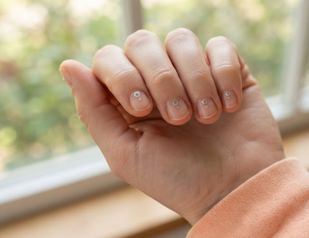 In this image, a full hand with extremely short round nails displays business nails with a bare nail and a silver floating dot, wearing a soft peach shirt.