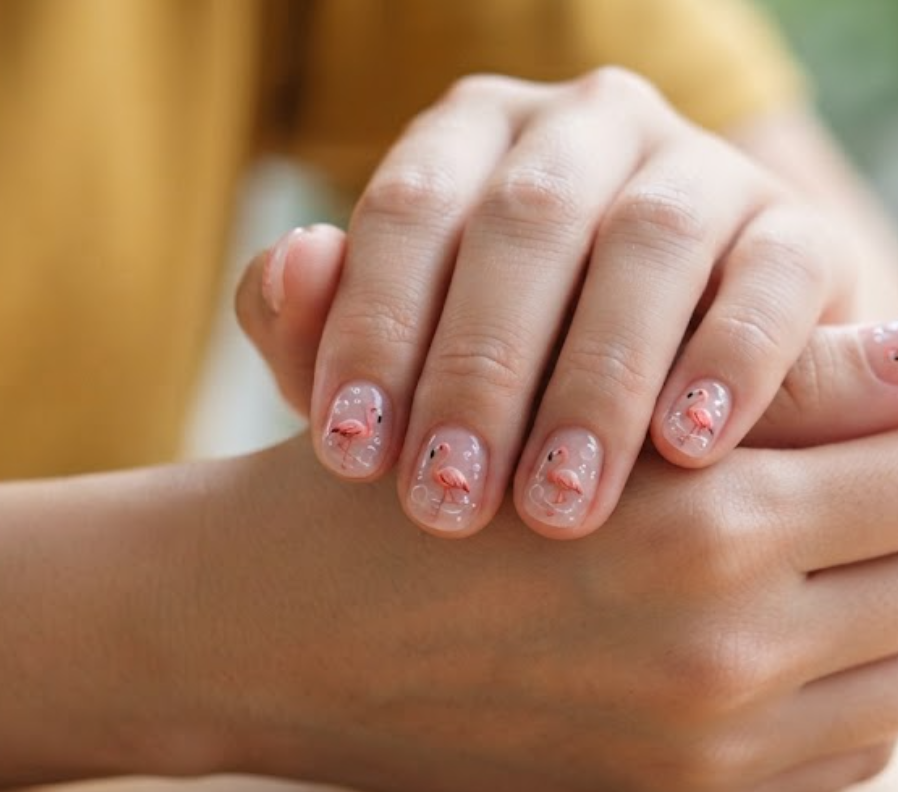 In this image, a woman in a mustard top presents short round nails with a translucent jelly pink base and floating flamingo.