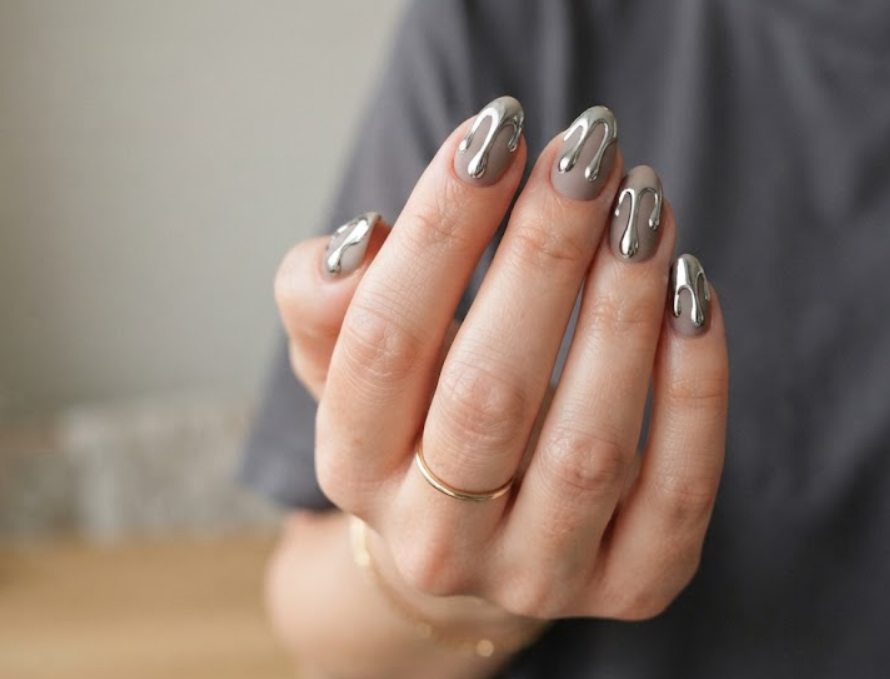 In this image, full hands with long almond nails showcase metallic silver melting drips over matte taupe against a clean indoor background.