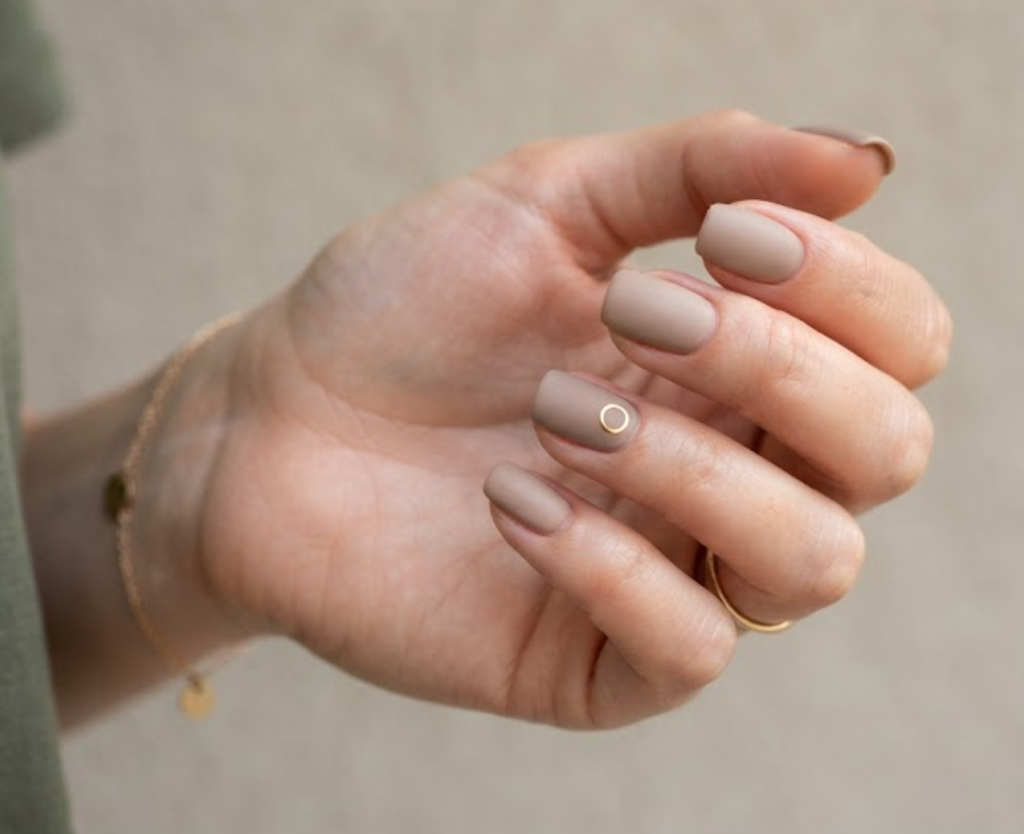 In this image, full hands with squoval nails feature tiny metallic ring decals on matte taupe against a neutral wall background.
