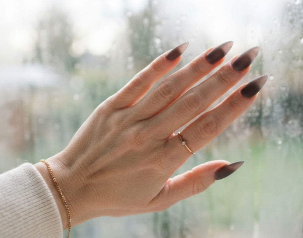 In this image, full hands with long stiletto nails show a frosted glass glaze over rich taupe against a window background with morning mist.