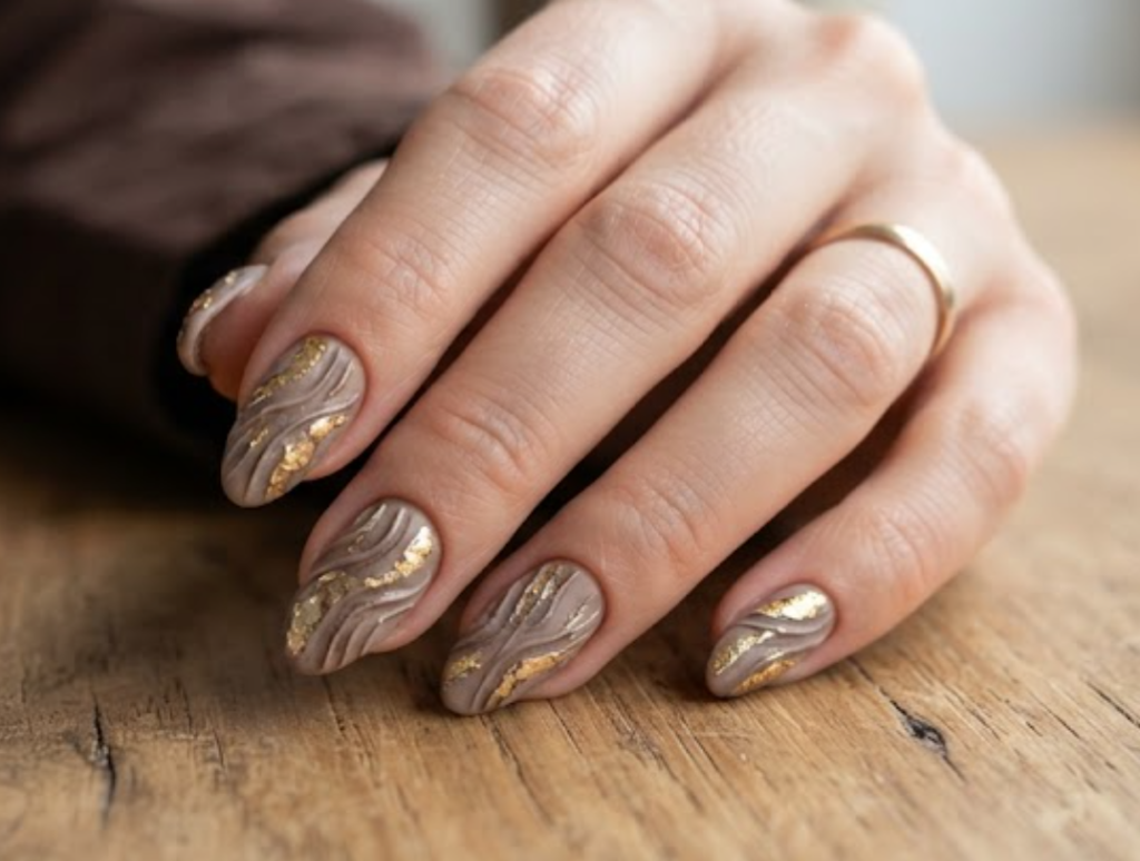 In this image, full hands with medium almond nails show textured grooves and gold layers on builder gel taupe against a wooden desk background.
