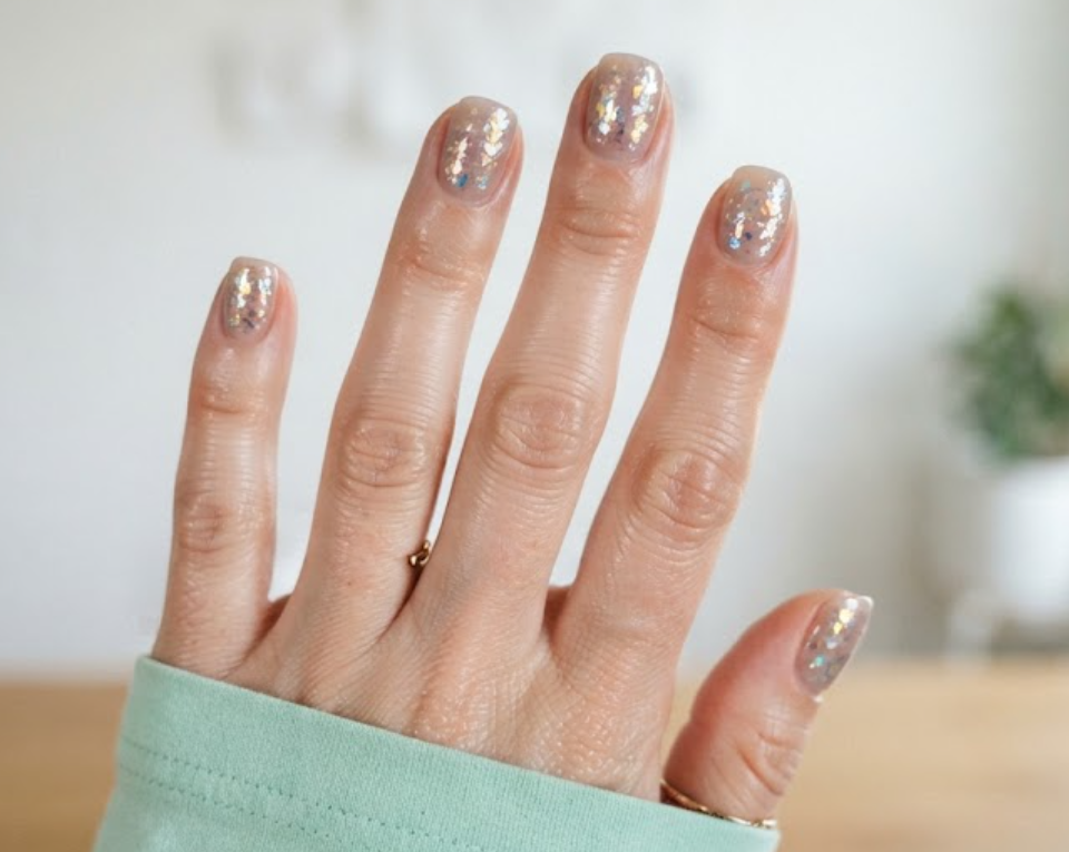 In this image, full hands with short squoval nails feature Mylar flakes under a milky taupe jelly coat against a bright room background.