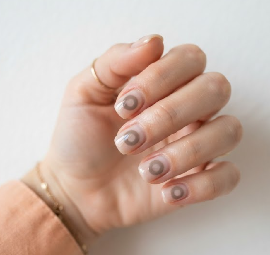 In this image, full hands with short square nails feature a floating taupe circular ring with negative space against a minimalist white background.
