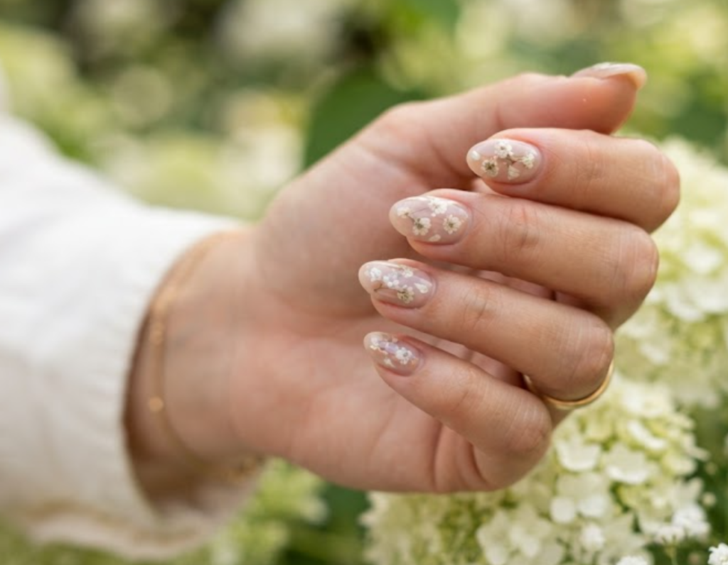 In this image, full hands with short almond nails showcase white baby's breath petals preserved inside a sheer taupe gel overlay against a floral background.