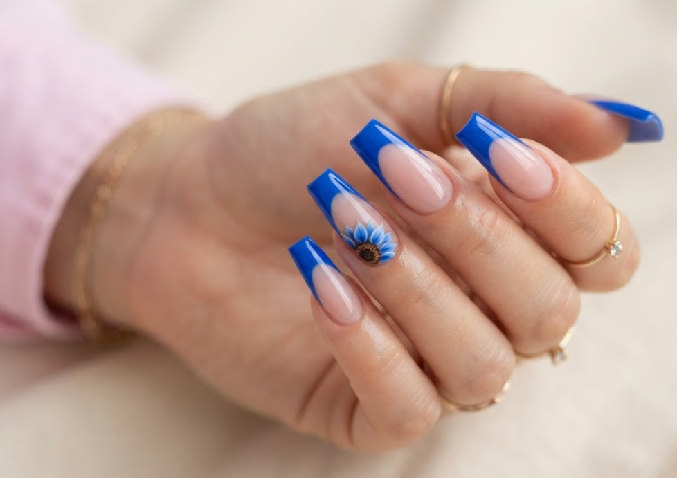 In this image, real hand nails highlight cobalt blue French tips with a sunflower accent and a pink shirt.