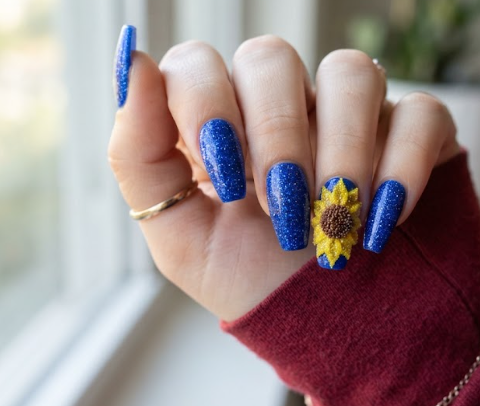 In this image, real hand nails showcase a royal blue glitter base with textured sunflower petals and a burgundy shirt.