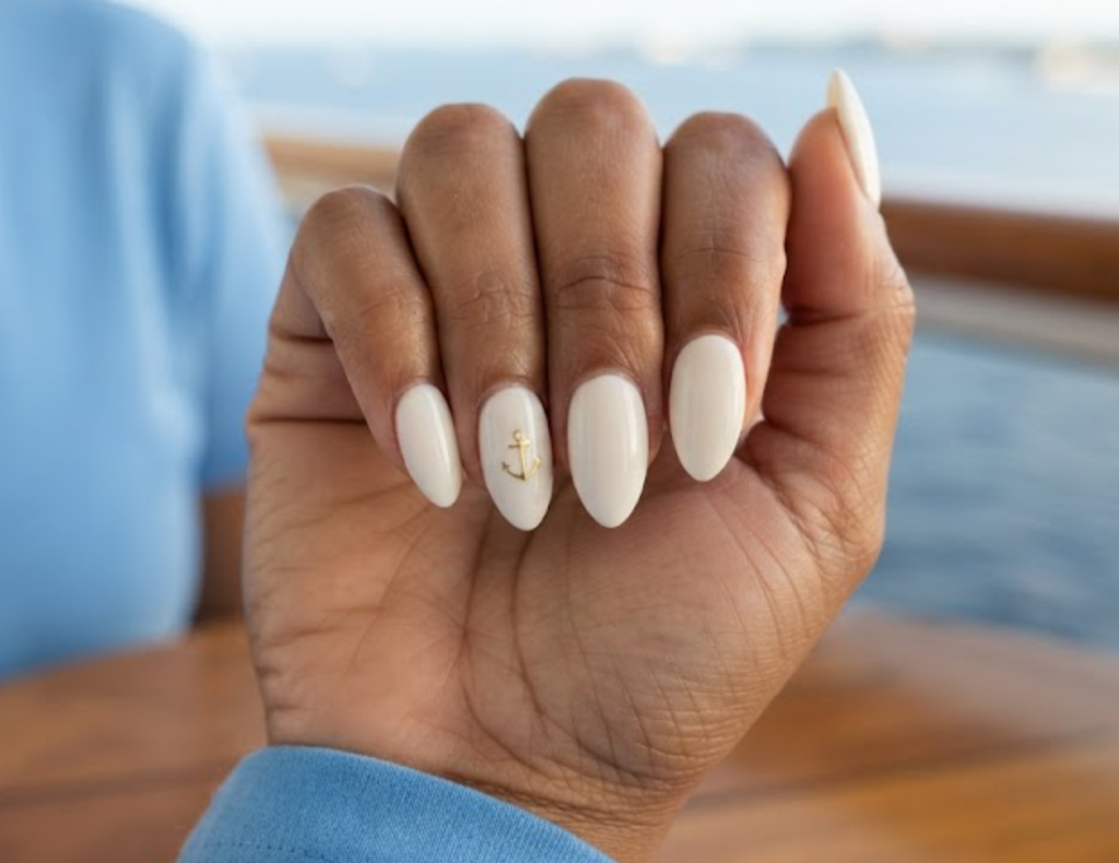 In this image, short almond nails feature ivory polish with a gold anchor decal. Realistic hands wearing a sky blue shirt appear against a blurred cruise balcony background.