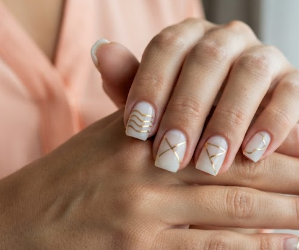 In this image, short square nails feature a milky base with thin gold geometric striping tape. Realistic hands wearing a soft peach blouse appear against a blurred window background.