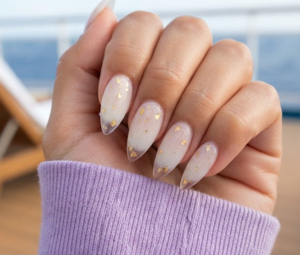 In this image, short stiletto nails feature a milky white to clear ombré with gold leaf flakes. Realistic hands wearing a lavender shirt appear against a blurred sun lounger background.