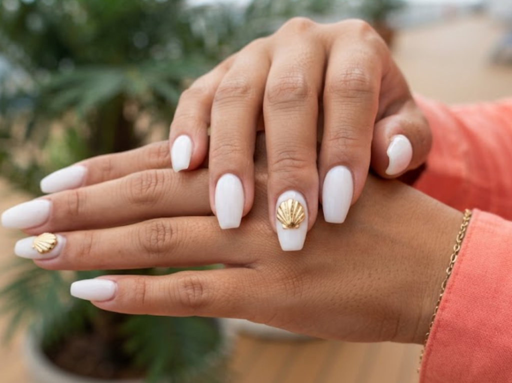 In this image, cruise wedding nails present short ballerina glossy white nails with a sculpted gold seashell, worn by hands in a coral shirt.
