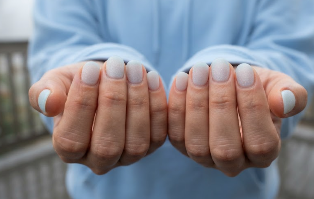 In this image, cruise wedding nails display short round icy frosted ocean shimmer nails, worn by hands in a powder blue hoodie.
