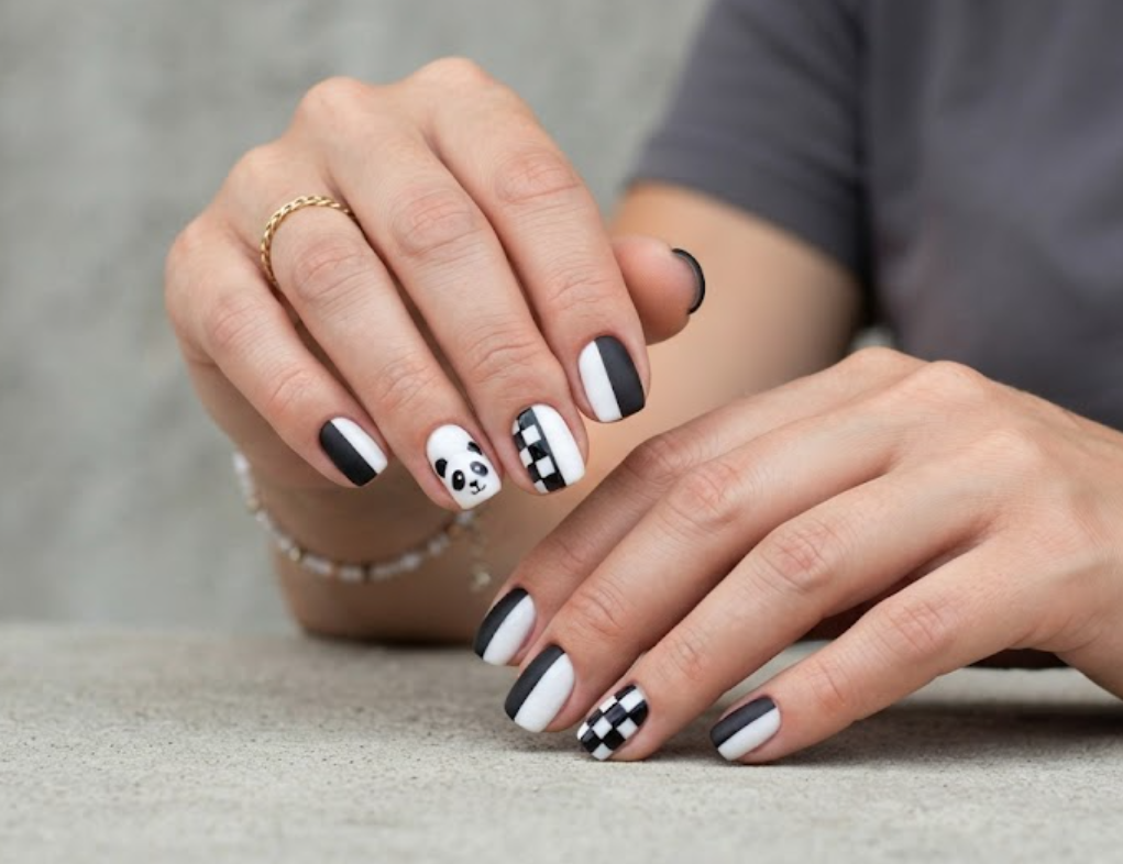 In this image, short squoval-shaped nails feature a modern vertical split base (matte black and glossy white). The white side of the accent nail shows half a panda face. The design includes color-blocking lines and a black-and-white checkerboard print. The hand wears a charcoal grey shirt.