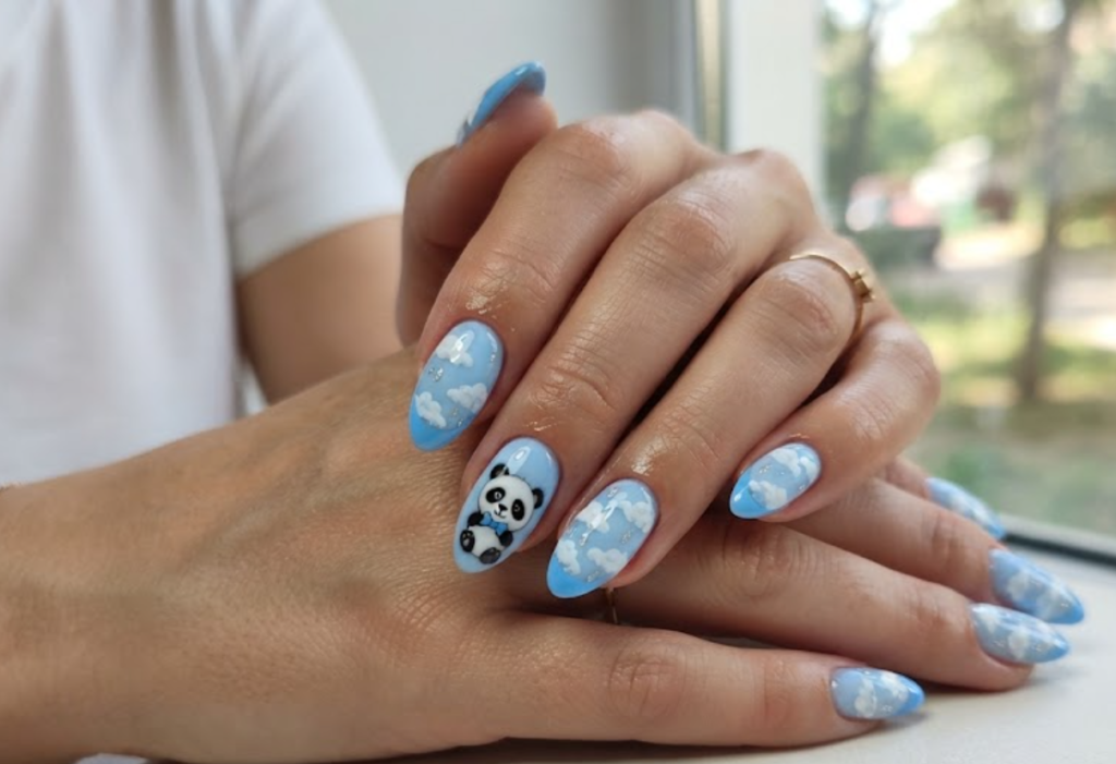 In this image, almond-shaped nails with a sky baby blue base are in sharp focus. One nail features a cute panda wearing a tiny blue bow tie. The design includes fluffy white clouds, silver raindrop accents, and solid blue French tips. The hand wears a solid white shirt.