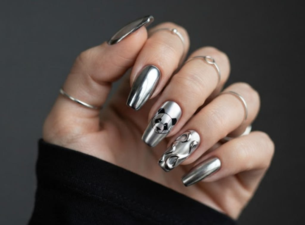 In this image, coffin-shaped nails with a metallic silver chrome finish are highlighted. The design includes a futuristic minimalist panda silhouette. Accents feature liquid metal swirls, metallic French borders, and mirror-finish solid nails. The hand wears a simple black shirt.