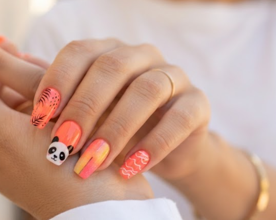 In this image, coffin-shaped nails with a bright coral orange base are in sharp focus. The accent nail has a cute panda face peeking from the corner. The design includes tropical leaf outlines, sunset color blocks, and white wave lines. The hand wears a white shirt against a sunlit wall.
