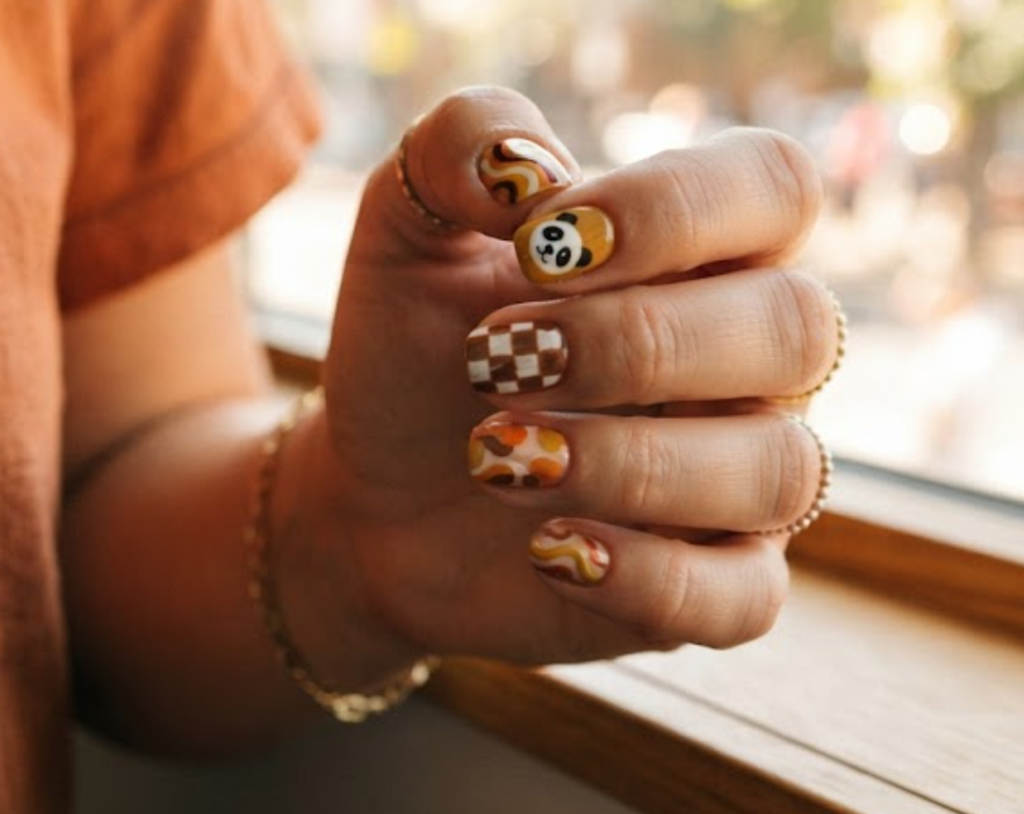 In this image, short square-shaped natural nails with a groovy mustard yellow base are shown. One nail displays a 70s retro-styled panda face. Others feature wavy 70s lines, brown-and-white checkers, and funky abstract shapes. The hand wears a burnt orange shirt against a blurred window.