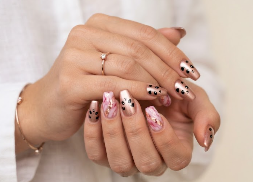 In this image, coffin-shaped nails with a metallic rose gold finish are highlighted. The design uses minimalist black lines for cute panda ears and eyes. Accents feature rose gold foil strips and glossy pink marbling. The hand wears a solid white blouse.