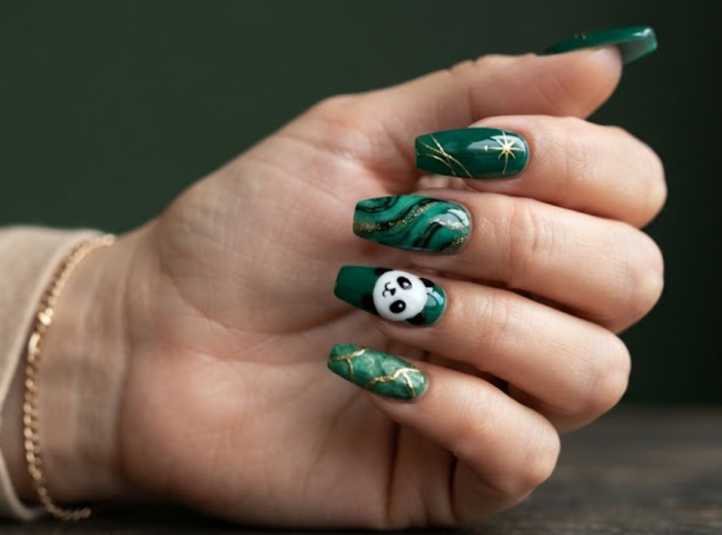 In this image, coffin-shaped nails with a rich glossy emerald green base are highlighted. The accent nail features a high-contrast white and black panda face. Others are styled with a gold-lined jade texture, marble swirls, and gold starbursts. The hand wears a beige shirt.
