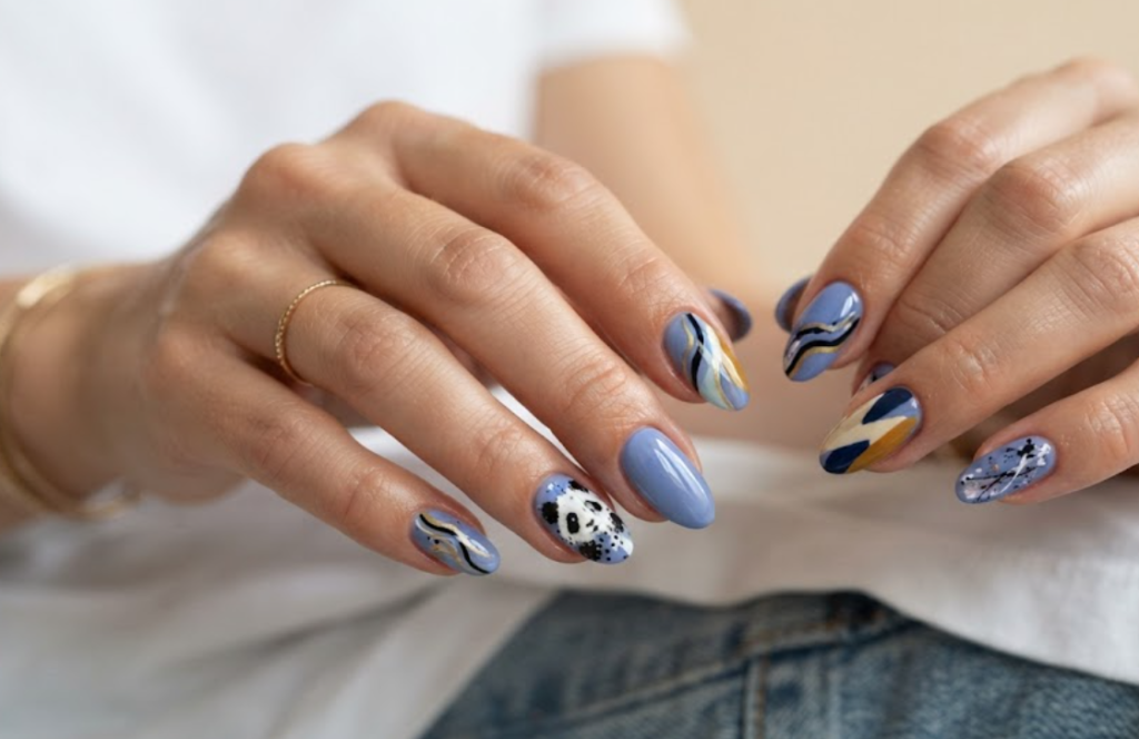 In this image, medium-length almond-shaped nails with a modern periwinkle blue base are highlighted. The accent nail displays an abstract brushstroke-style panda face. Others feature modern color blocks, wavy artistic lines, and minimalist paint splatters. The hand wears a white shirt.