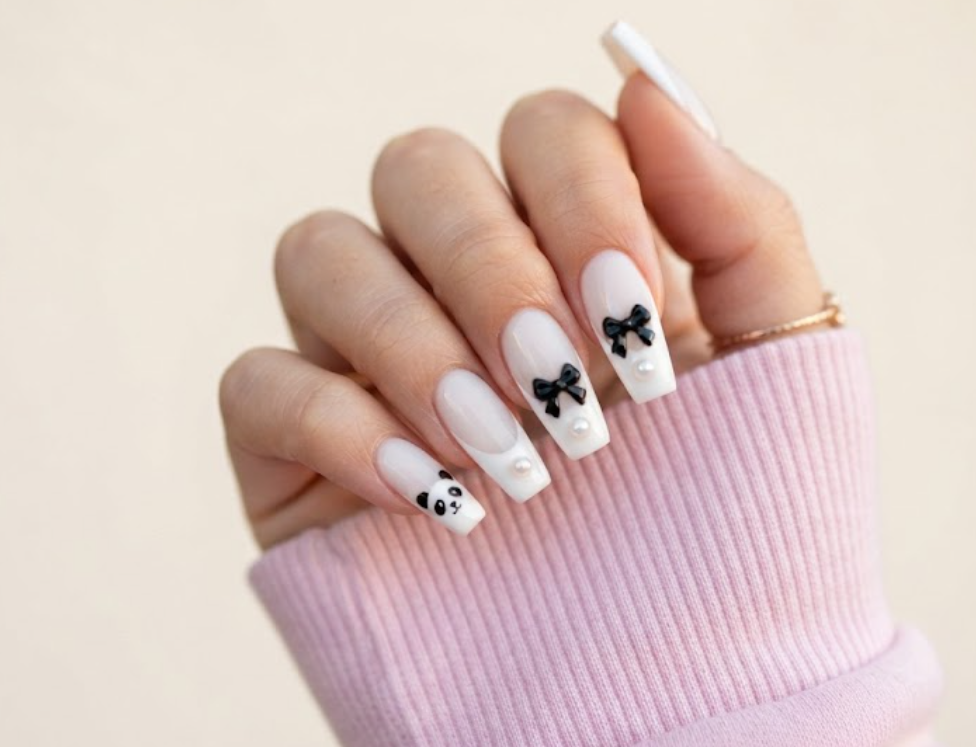 In this image, coffin-shaped nails with a translucent milky white base are shown. The accent nail features a classic French tip with a tiny panda face in the corner. Others have classic tips with tiny black bows, pearl studs, and clean negative space. The hand wears a pastel pink shirt.