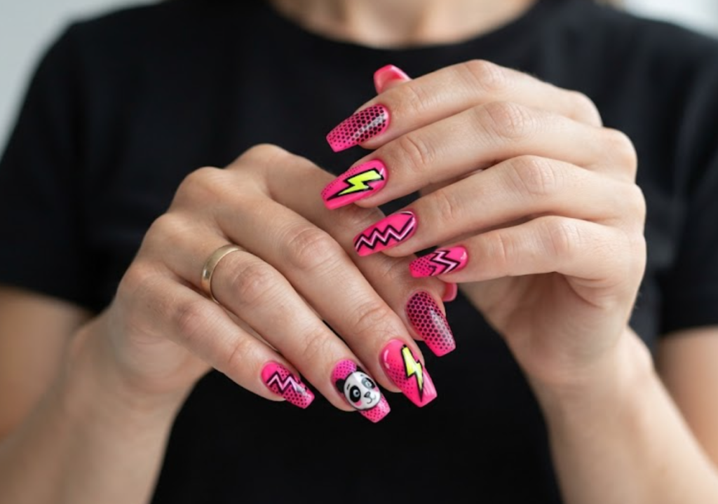 In this image, coffin-shaped nails with a vibrant hot pink base are highlighted. The accent nail features a pop-art style comic panda face with bold outlines. Others are decorated with black halftone dots, neon yellow lightning bolts, and bold zig-zag lines. The hand wears a black t-shirt.