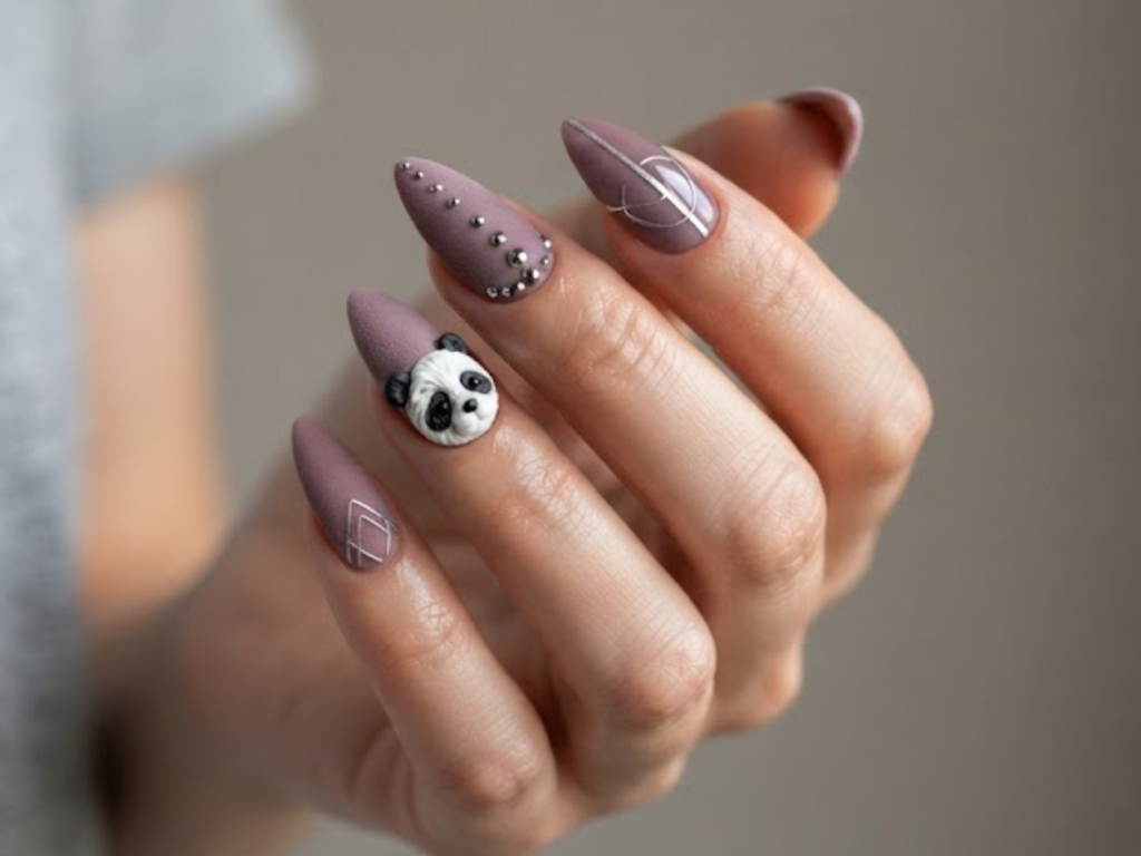 In this image, medium-length stiletto nails with a dusty mauve matte velvet finish are in sharp focus. The accent nail features a 3D textured panda face. Others display minimalist silver wire-art lines, matte-and-glossy split nails, and tiny metallic studs. The hand wears a grey t-shirt.