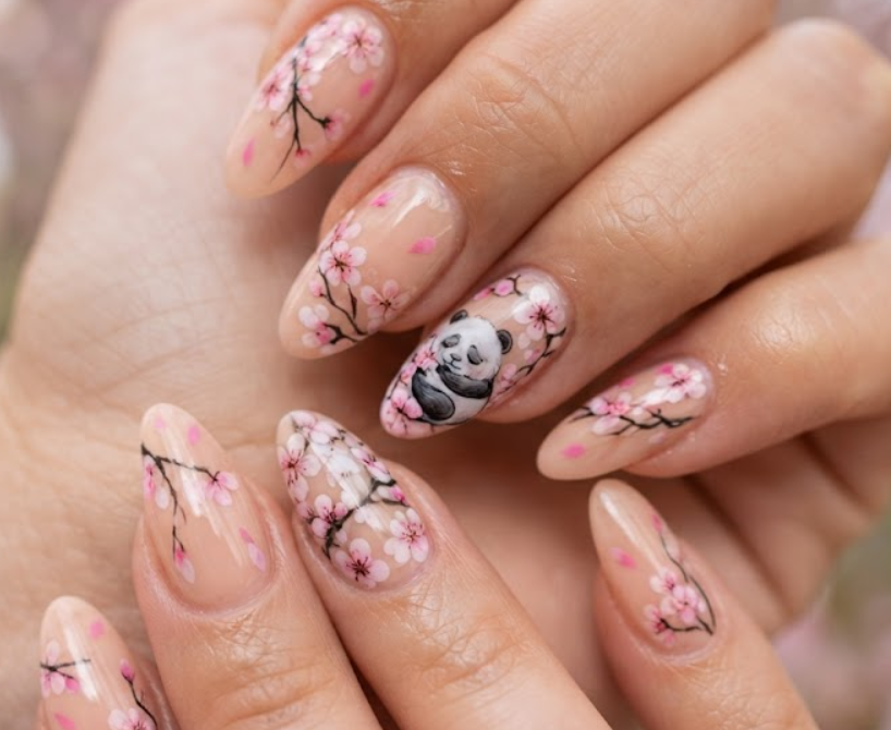 In this image, medium-length stiletto nails with a soft translucent peach base are in sharp focus. The accent nail features a sleeping panda nestled among cherry blossoms. Others display falling pink cherry blossom petals, delicate branch lines, and translucent nude glossy coats. The hand wears a light pink blouse.
