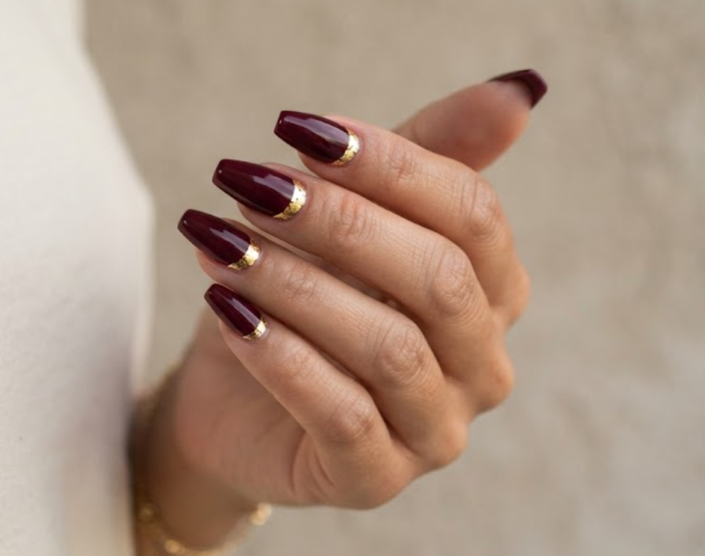 In this image, a real hand with deep burgundy coffin nails featuring cuticle gold foil crescents is shown in a close-up photo, wearing a simple solid cream colored shirt, simple neutral wall background, natural lighting, realistic photography.