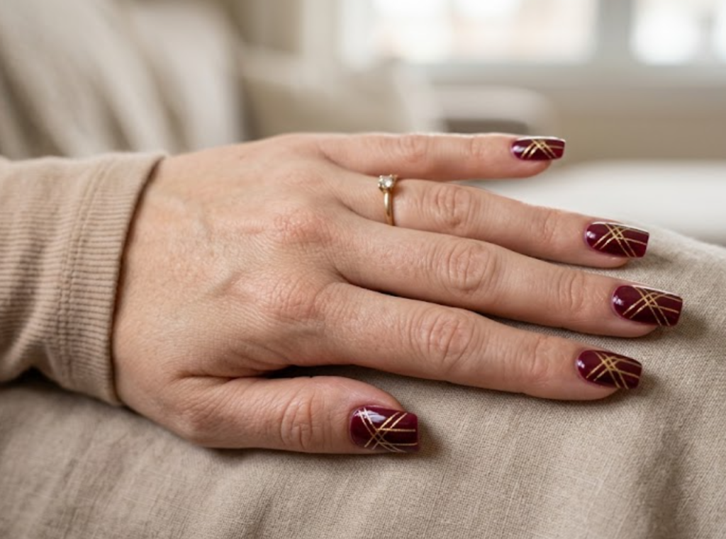 In this image, a real hand with structured square wine-red nails featuring interlocking gold geometric lines is shown in a close-up photo, wearing a simple solid warm beige shirt, simple soft indoor background, natural lighting, realistic photography.