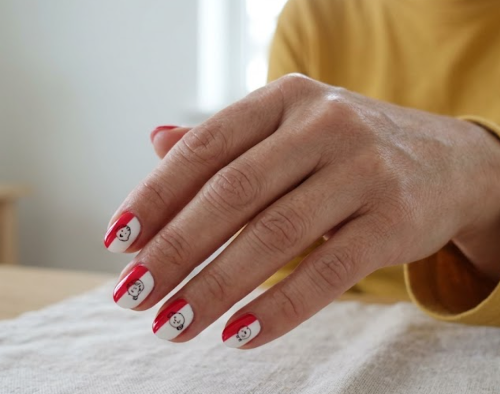 In this image, a real hand with oval red and white split nails featuring miniature character graphics is shown in a close-up photo, wearing a simple solid mustard yellow shirt, simple minimal room background, natural lighting, realistic photography.