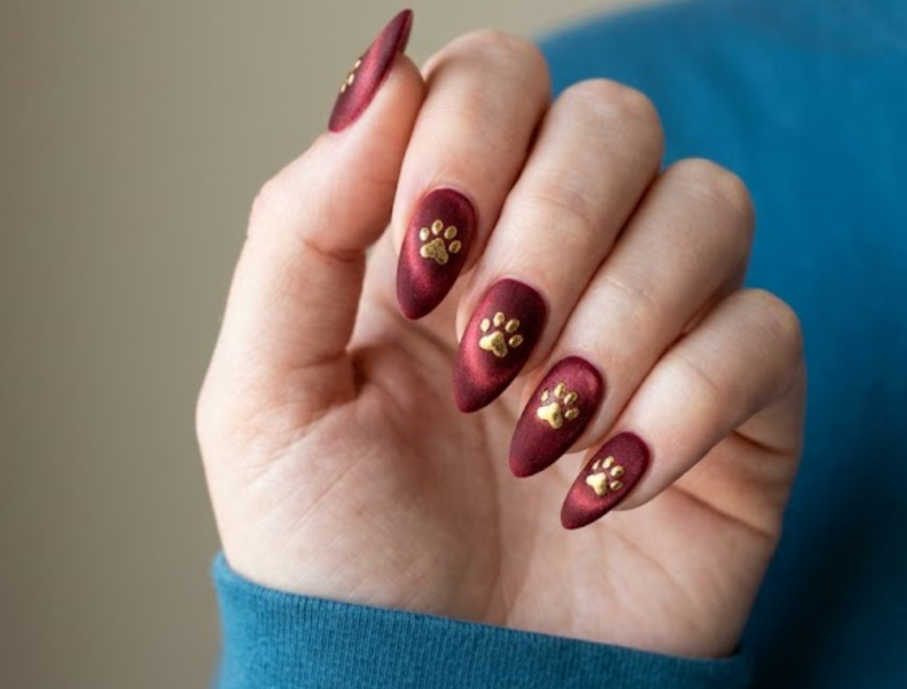 In this image, a real hand with stiletto velvet red cat-eye nails featuring embossed golden animal paw prints is shown in a close-up photo, wearing a simple solid teal blue shirt, simple plain background, natural lighting, realistic photography.