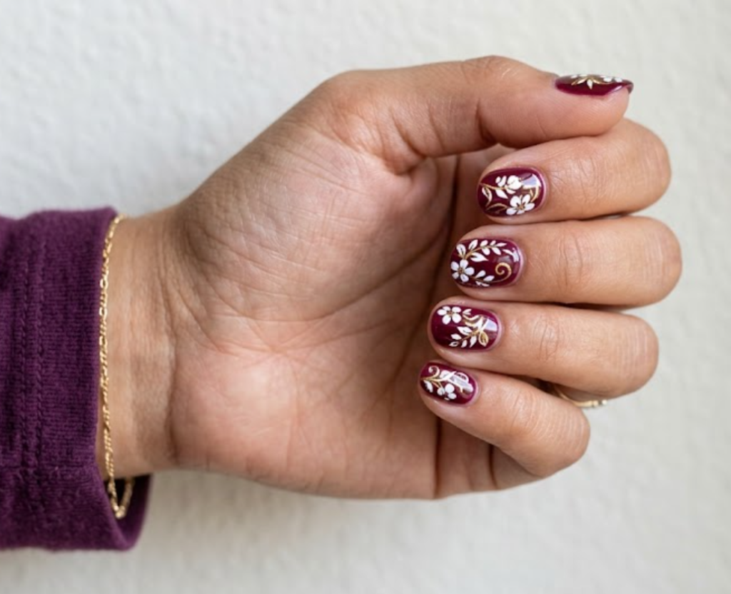In this image, a real hand with short round burgundy nails featuring white silhouette florals and gold outlines is shown in a close-up photo, wearing a simple solid plum purple shirt, simple neutral wall background, natural lighting, realistic photography.