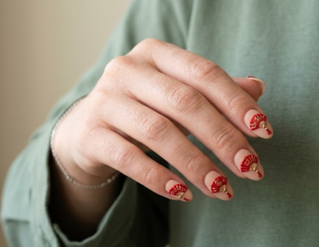 In this image, a real hand with deep red coffin nails paired with tiny gold coin charms and studs is shown in a close-up photo, wearing a simple solid crimson red shirt, simple clean indoor background, natural lighting, realistic photography.