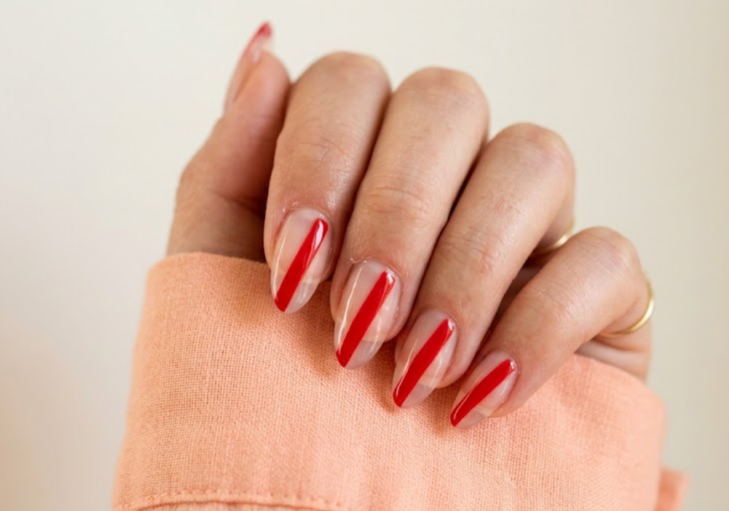 imageIn this close-up photo, Chinese New Year nails featuring transparent almond tips with bold red negative space diagonal stripes are shown on a real hand wearing a soft peach shirt.