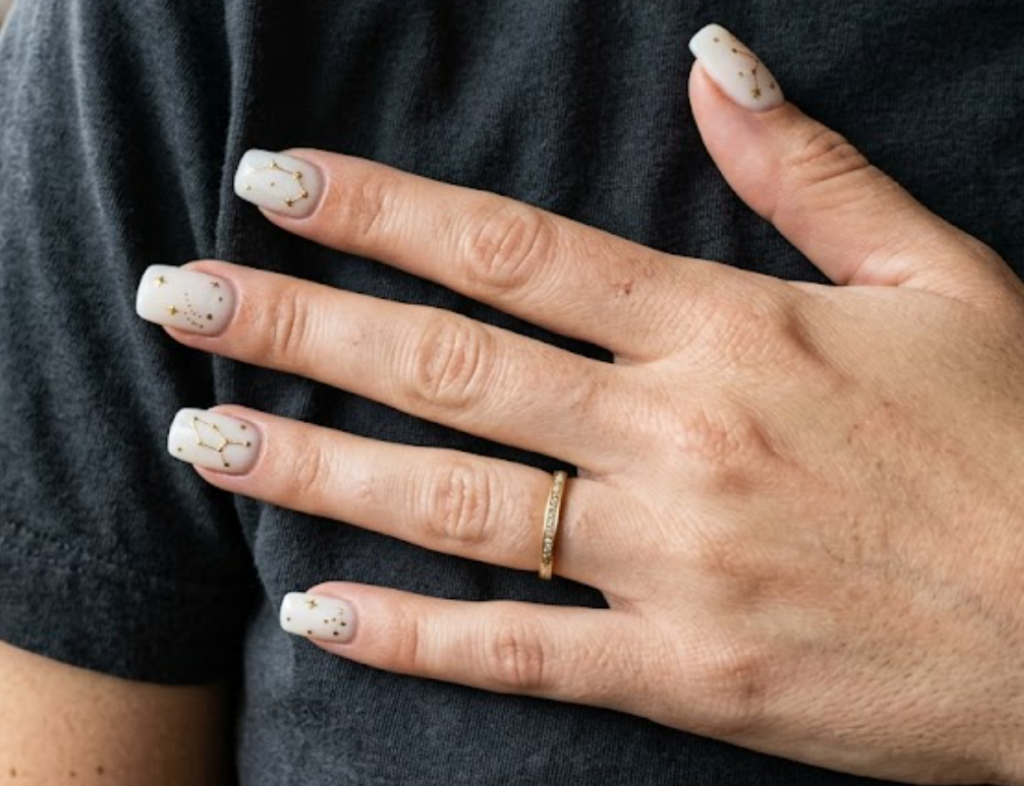 In this close-up photo, Chinese New Year nails featuring milky white square tips with micro gold constellations and dots are shown on a real hand wearing a charcoal shirt.