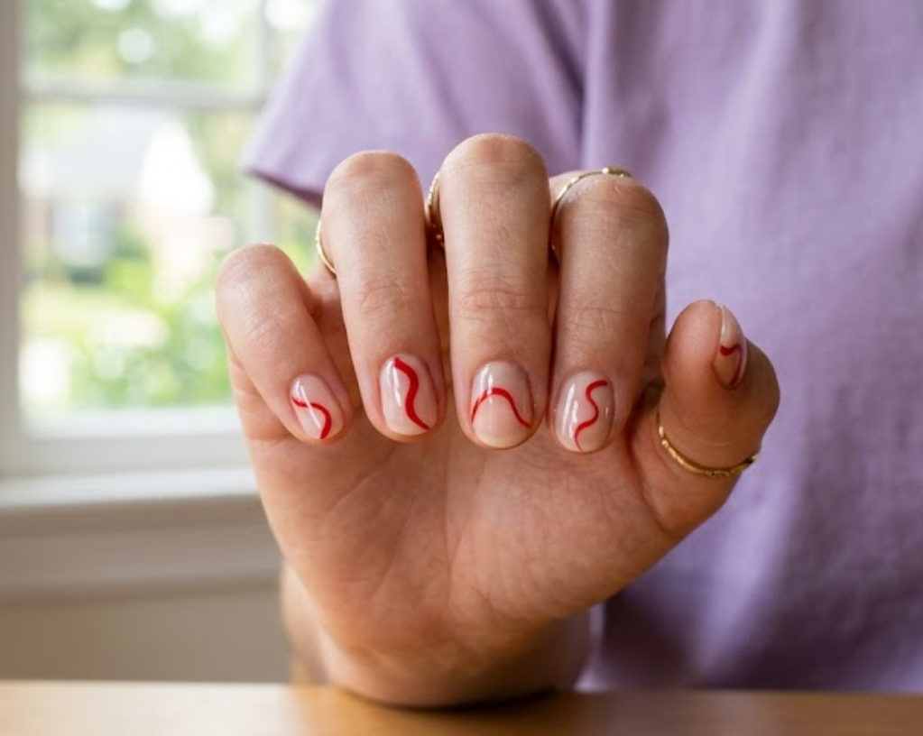 In this close-up photo, Chinese New Year nails featuring short round glossy clear tips with abstract curved red wave lines are shown on a real hand wearing a lavender shirt.